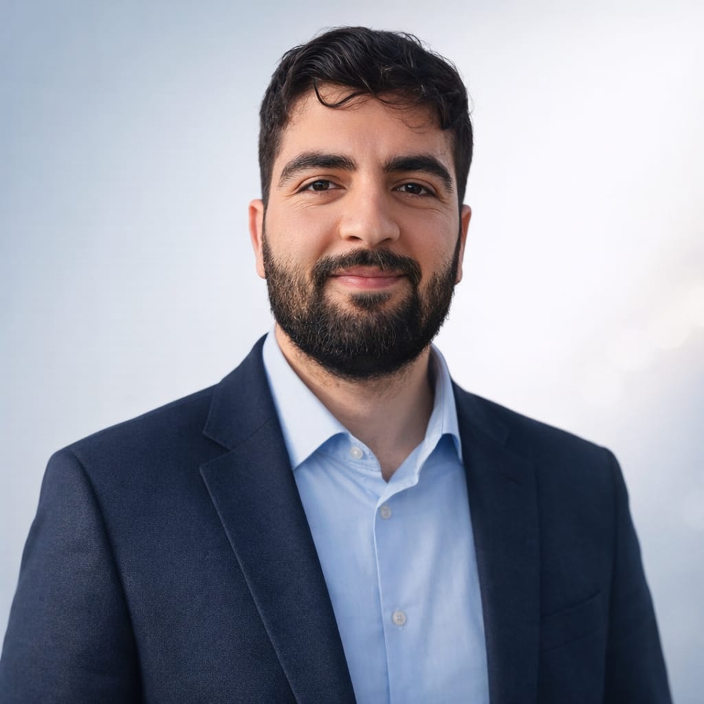 A professional headshot of a man with dark hair and a beard, wearing a navy blazer and light blue shirt, smiling slightly against a soft, neutral background.